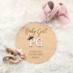 Round Plywood Baby Announcement Sign with White 3D Acrylic Initials, Accented by a Rose Gold Mirror Crown and Engraved Text, Positioned on a Shaggy White Rug, Flanked by Baby Shoes and a Teether in the Background.