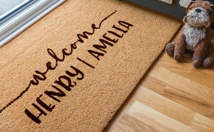 Personalised door mats: 900x600mm brown coir mat with black UV printed 'welcome' and custom names, displayed on a timber floorboard at the front doorstep.